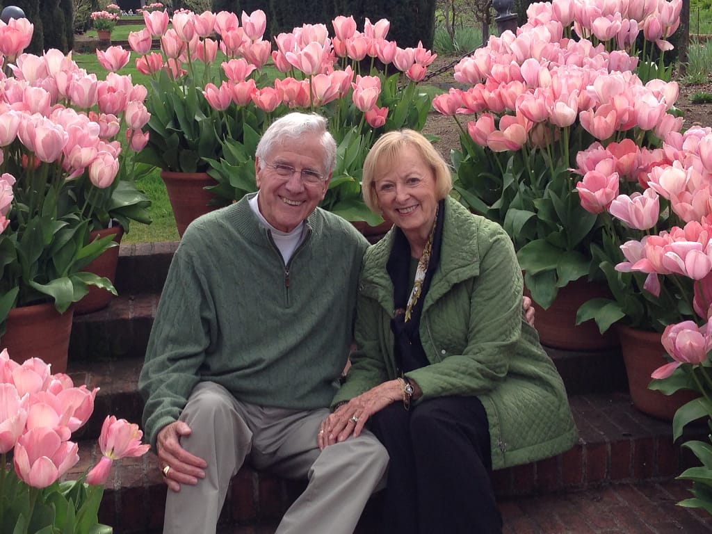 Barbara Little's Parents seated and surrounded by pink flowers.