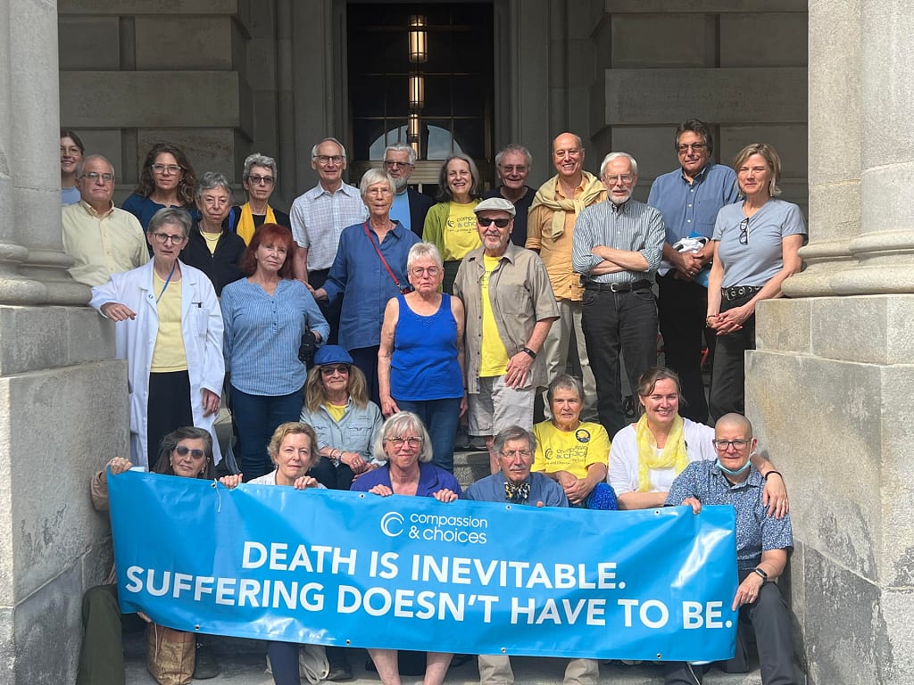 Group of protestors outside of NY State House holding a banner saying "Death is Inevitable. Suffering Doesn't Have to Be."