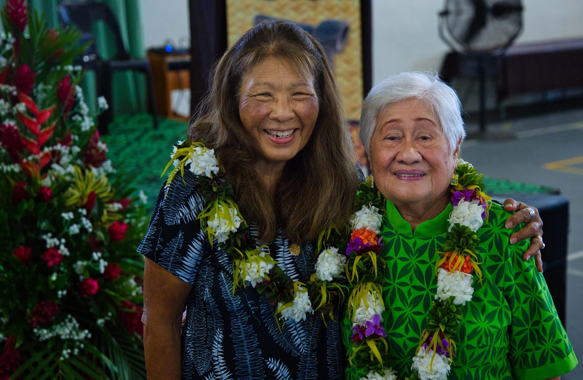 Two women wearing leis and floral shirts, smiling at the camera.