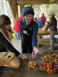 Alice Smith holds an exotic looking mushroom in her hand with several other varieties around her. She is displaying it to a young woman.