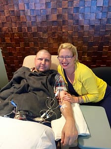 White man sitting in a cream colored chair with tubes coming from his arm, covered in blankets with a blonde woman with glasses and a yellow cardigan smiling next to him.