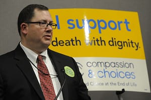 tim appleton stand in front of a sign that reads "I Support Death with Dignity"