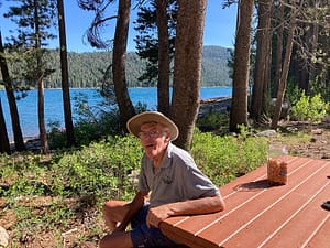 Barbara’s husband, Dick Kuehner sits at a picnic table in front of a tree-lined lake