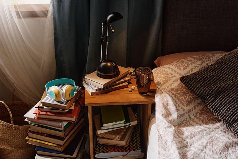A bedside table with a lamp, headphones and stacks of books.